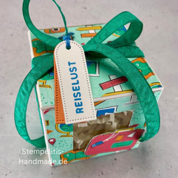 Handdekorierte Fensterschachtel mit Reisethema und grünem Band, Detailansicht des Sichtfensters mit Füllmaterial, Koffer-Motiv auf der Front der Schachtel, Oberseite mit Geschenkanhänger „Reiselust“, Seitliche Ansicht der Box mit Weltkartenmotiv, Geöffnete Box mit Blick auf das Innere, Fensterschachtel als kreative Geschenkverpackung für Reisegutscheine